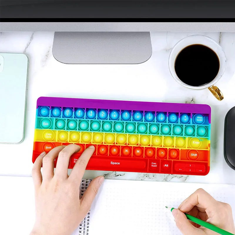 Hands popping the rainbow pop keyboard at a desk next to a notebook